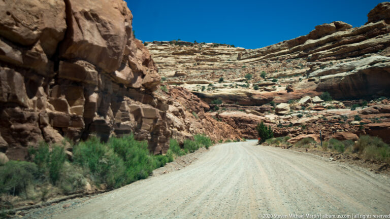 A pale gravel road curves between close sandstone walls and sparse canyon-floor shrubs.
