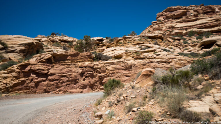 A paved road curves around a layered sandstone slope scattered with loose slabs and shrubs.