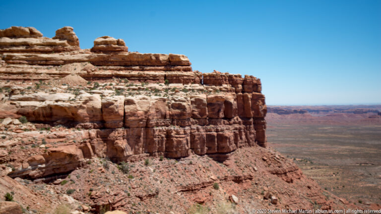 A layered sandstone cliff drops to a broad desert plain with mesas on the horizon.