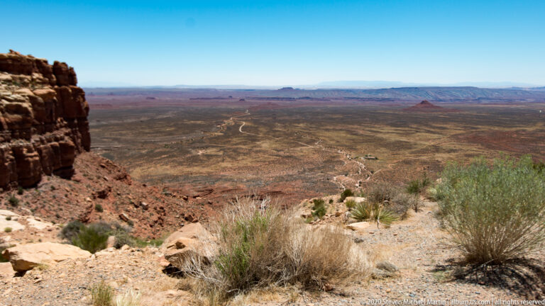 A rocky rim overlooks sparse desert scrub, an open plateau, and distant blue mesas.