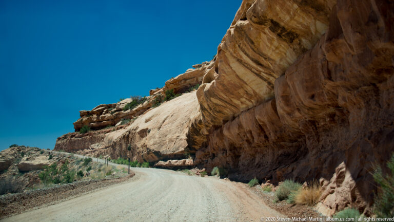A gravel road follows a stone barrier beneath a steeply tilted sandstone overhang.