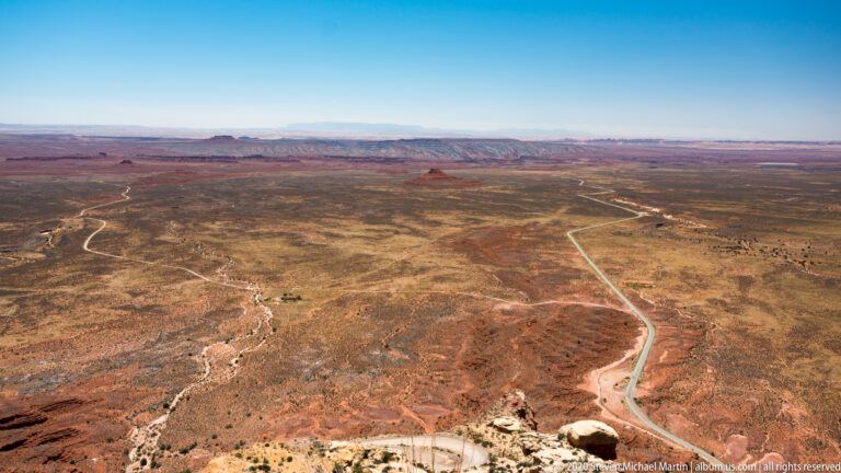 Pale roads wind across a broad red-brown plateau toward distant mesas and blue ridges.