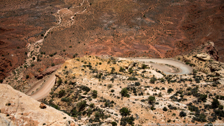 A narrow road forms a hairpin around a pale outcrop on a red, shrub-dotted slope.