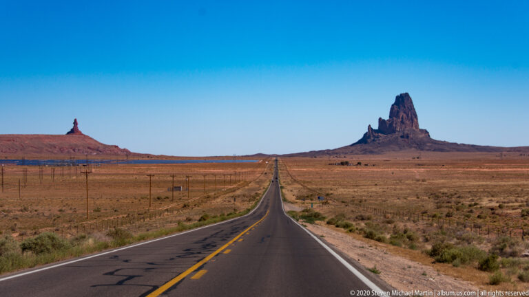 A straight two-lane road leads toward a tall desert monolith between distant buttes.