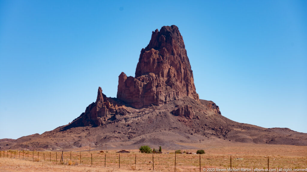 A tall red monolith with an uneven pinnacled summit rises behind a desert fence.