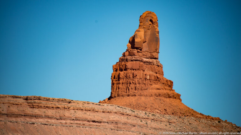 A narrow red pinnacle rises from a broad, horizontally layered mesa base.