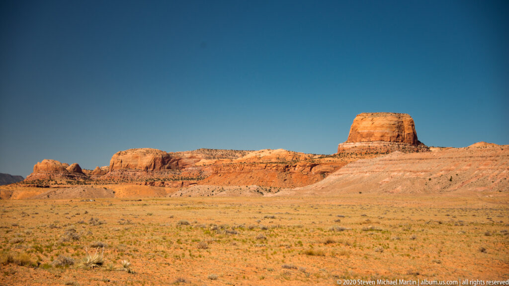 A broad flat-topped mesa rises above a grassy, shrub-dotted open desert plain.