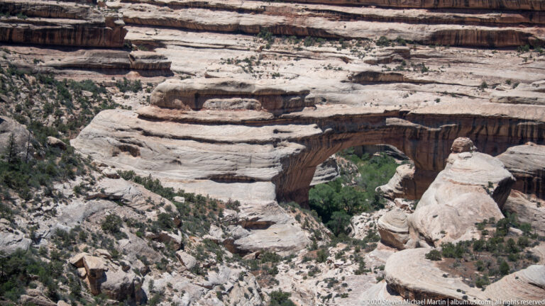 A broad sandstone bridge crosses a green, tree-filled ravine within pale layered cliffs.