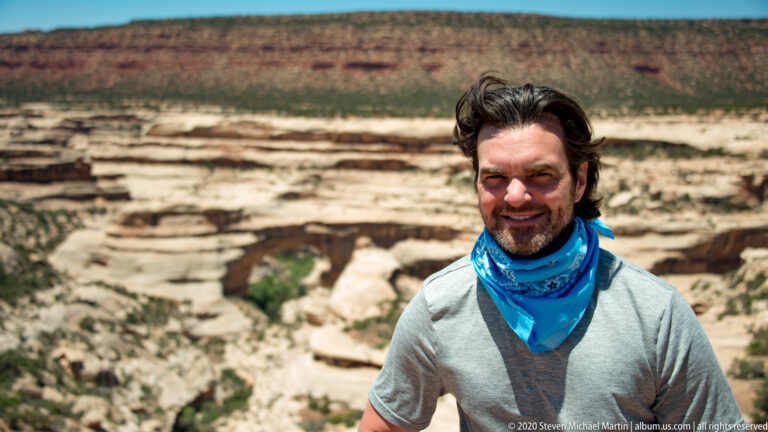 A smiling man in a gray shirt and blue neck gaiter stands before a canyon bridge.