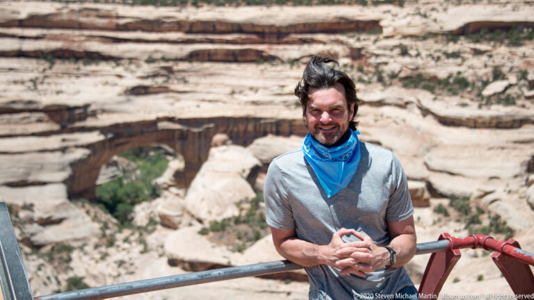 A man leans on a red overlook railing with a sandstone bridge behind him.