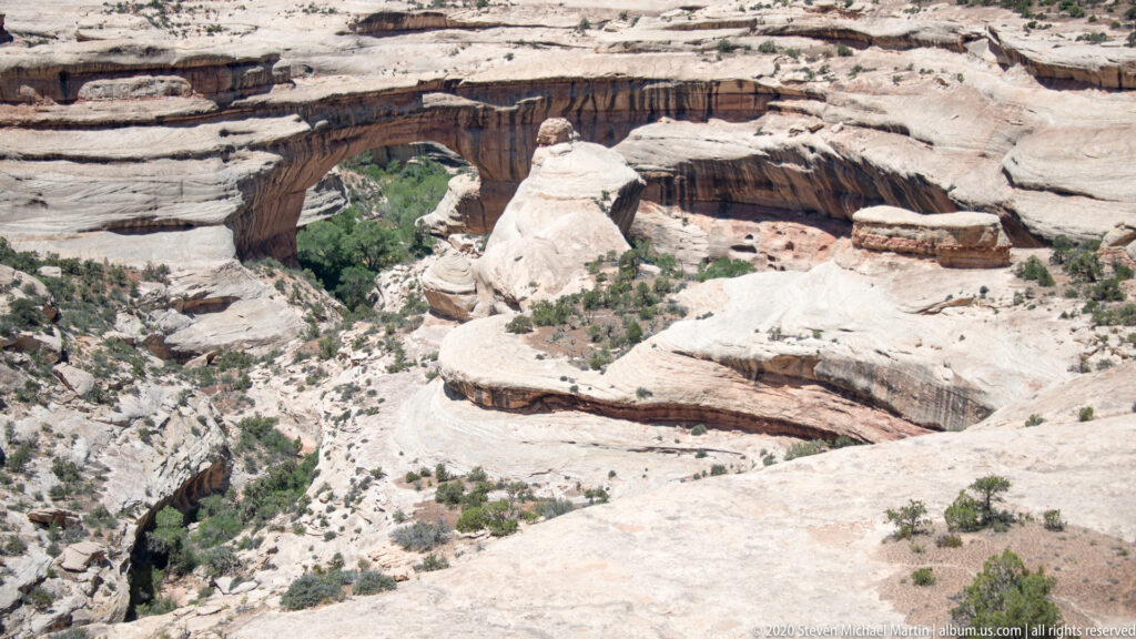 A sandstone bridge crosses a broad pale canyon above green vegetation and rounded ledges.