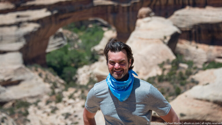 A smiling man in a gray shirt and blue neck gaiter stands before a blurred bridge and canyon.