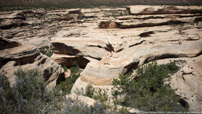 Layered pale sandstone ledges and shallow channels descend toward shrubs below a mesa.