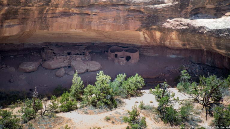Stone rooms with small dark openings sit beneath a broad sandstone alcove.