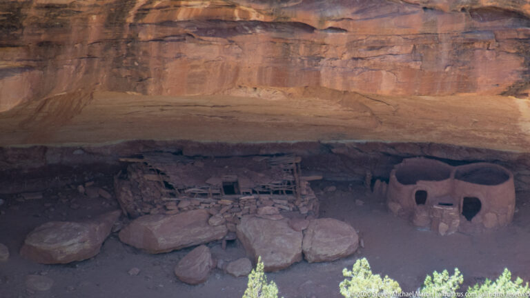 Several low stone rooms and boulders occupy the shaded back of a broad sandstone alcove.