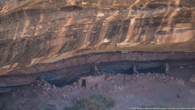 Low masonry remains line the shaded base of a dark-streaked sandstone overhang.