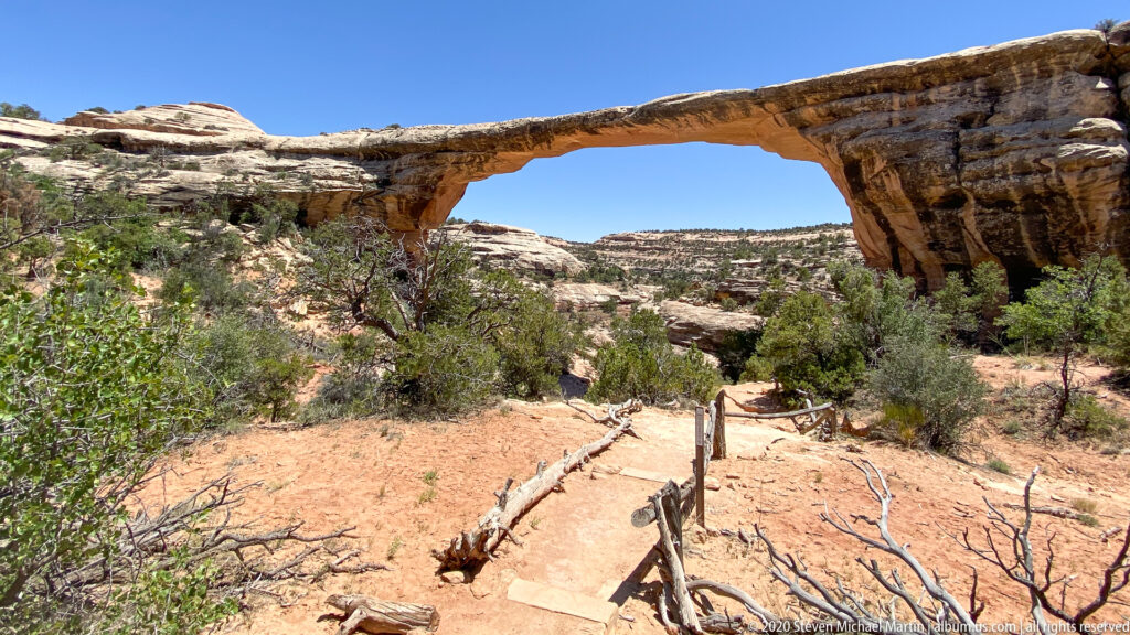 A sandstone bridge arches over a sandy visitor path edged by a low wooden rail.