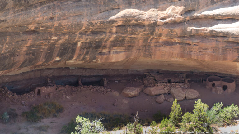Low stone structures sit in shadow beneath a long reddish sandstone overhang.