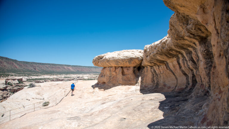 A visitor walks along a handrailed rock path toward a broad sandstone bridge beneath a cliff.