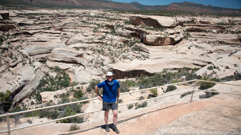 A man stands beside an overlook rail above pale slickrock benches and distant red mesas.