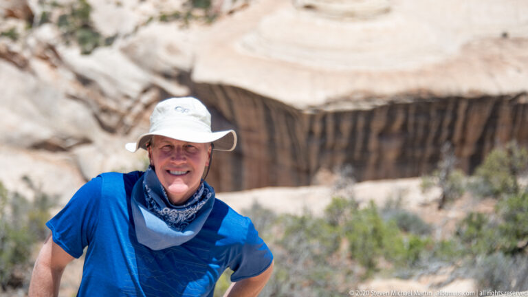 A smiling visitor in a blue shirt and sun hat stands before a blurred sandstone bridge opening.