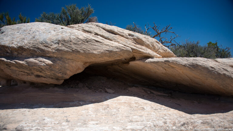 A pale sandstone shelf projects over a dark shallow recess, with shrubs beyond the rock.