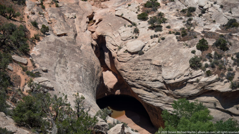 A deep cleft between sandstone walls descends to a dark opening among ledges and shrubs.