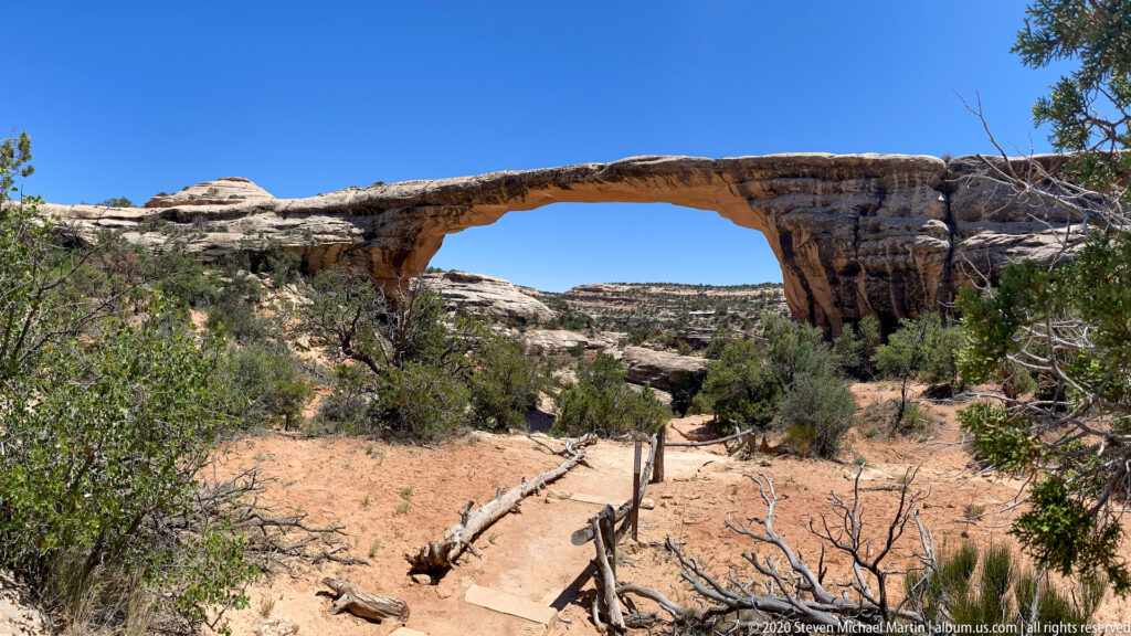 A long sandstone bridge spans a sandy trail surrounded by shrubs and dry branches.