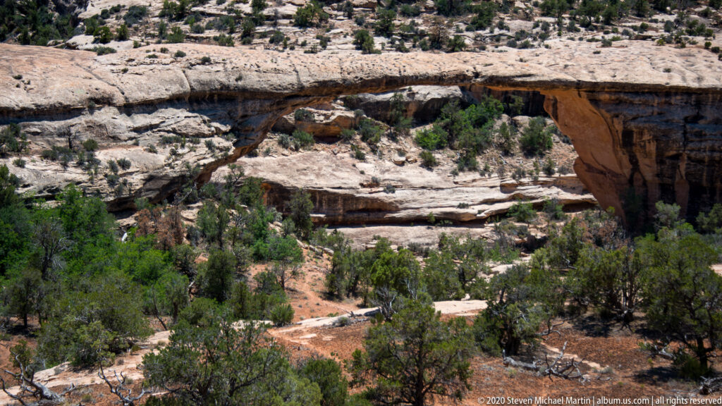 A sandstone bridge crosses above a shaded ravine with trees and a sandy wash below.