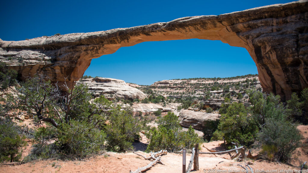 A high sandstone bridge frames pale canyon ledges, shrubs, and a sandy path through its opening.