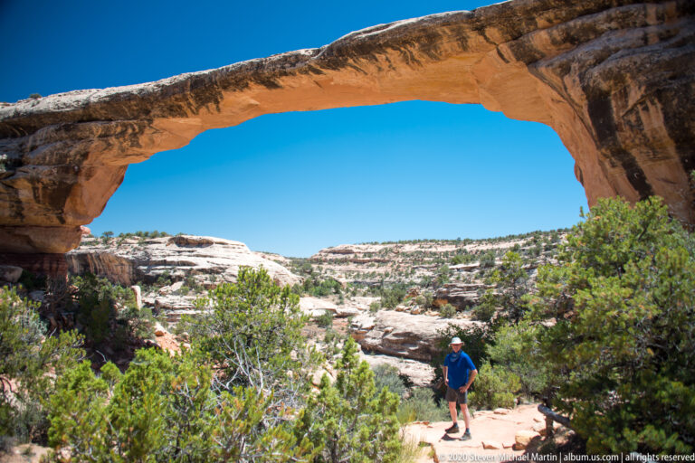 A hiker stands beneath a massive sandstone bridge above a sandy trail and canyon shrubs.