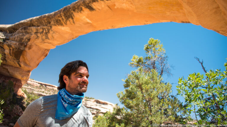 A man in a gray shirt and blue scarf stands beneath an orange sandstone bridge opening.
