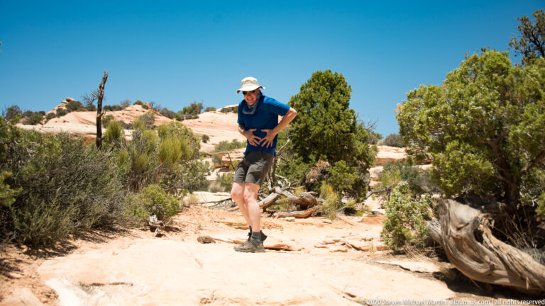 A hiker pauses on pale sandstone among juniper shrubs and fallen branches.