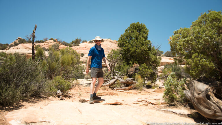 A hiker walks toward the camera on a sandy slickrock path between shrubs and fallen wood.