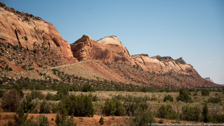 A red layered escarpment stretches above juniper-covered ground beneath a clear blue sky.
