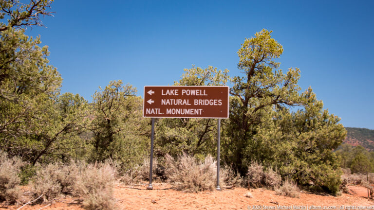 A brown sign points toward Lake Powell and Natural Bridges National Monument among junipers and dry brush.