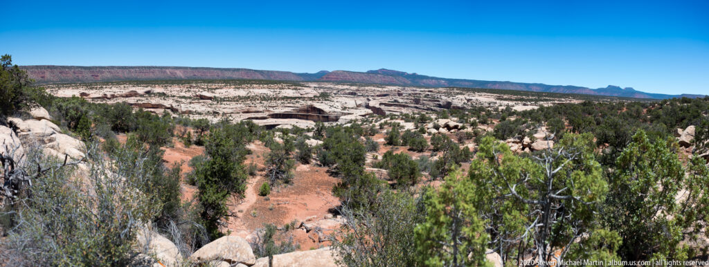 A sandy trail descends through junipers toward pale canyon rock and distant red mesas.