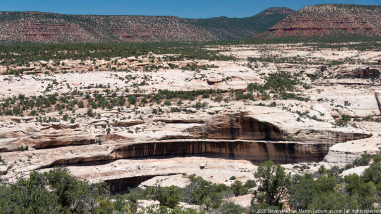 A pale sandstone ledge overhangs a dark recess above trees and distant red mesas.