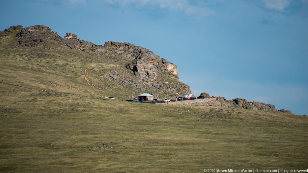 A small roadside building and parked vehicles sit below a broken alpine rock ridge.