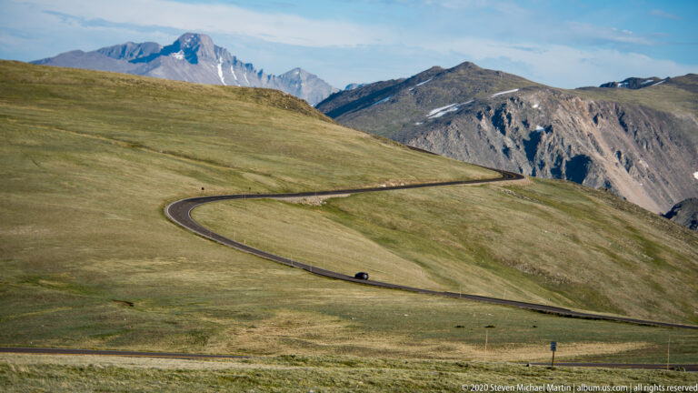 A winding paved road traverses an open alpine hillside beneath distant snow-patched mountain peaks.