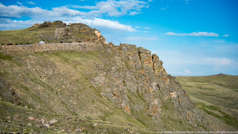 A blocky gray and rust-colored outcrop rises from an open alpine grass slope.