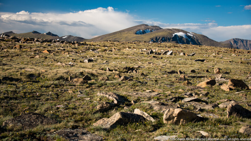 Rocky alpine tundra with short grasses opens toward distant mountain slopes and snow patches.