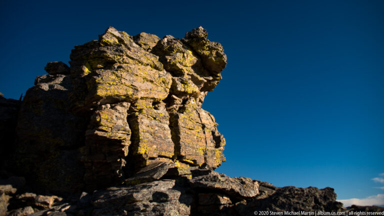 Tall rough rock spire mottled with yellow-green lichen rises against blue sky.