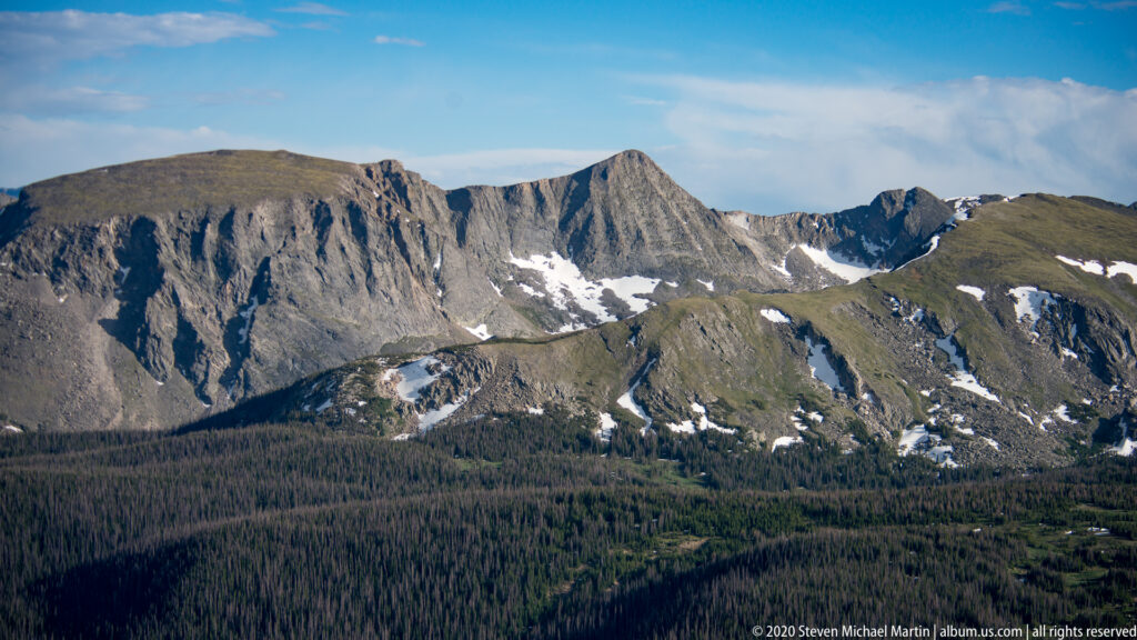 Snow patches cling to jagged gray peaks above a dark evergreen forest.