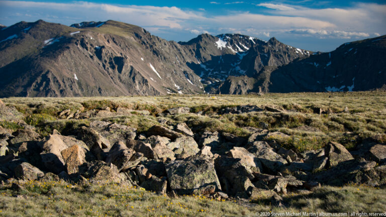 Boulders and low flowers fill a high meadow beneath steep dark mountains with snow patches.