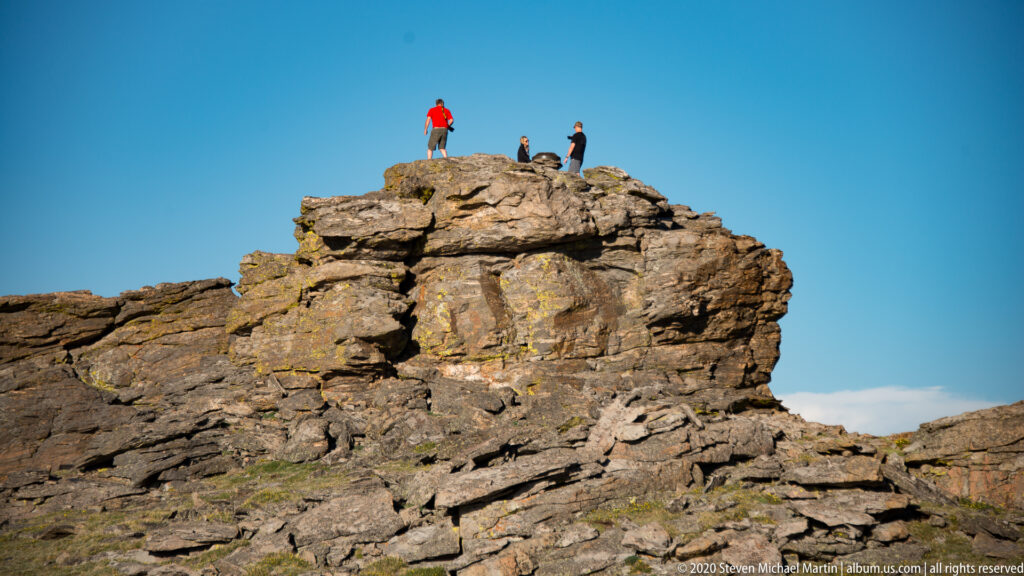 Three hikers, including one in a red-orange top, stand on an exposed granite ledge.