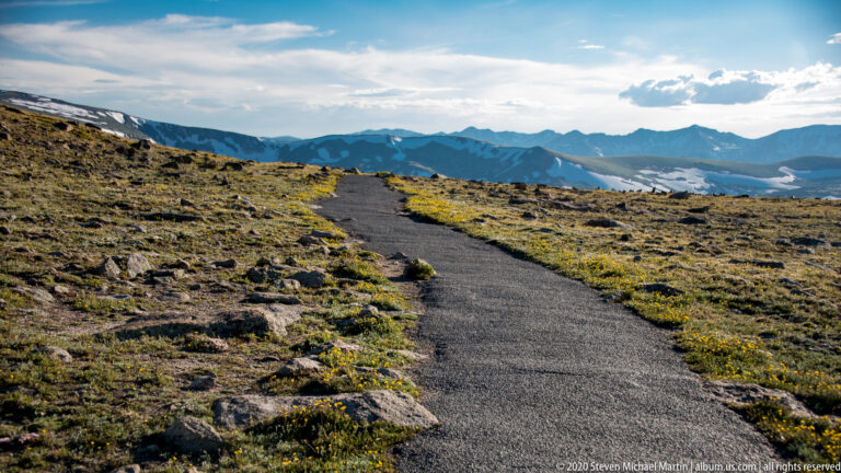 A narrow trail curves across low alpine vegetation toward rugged distant mountains.