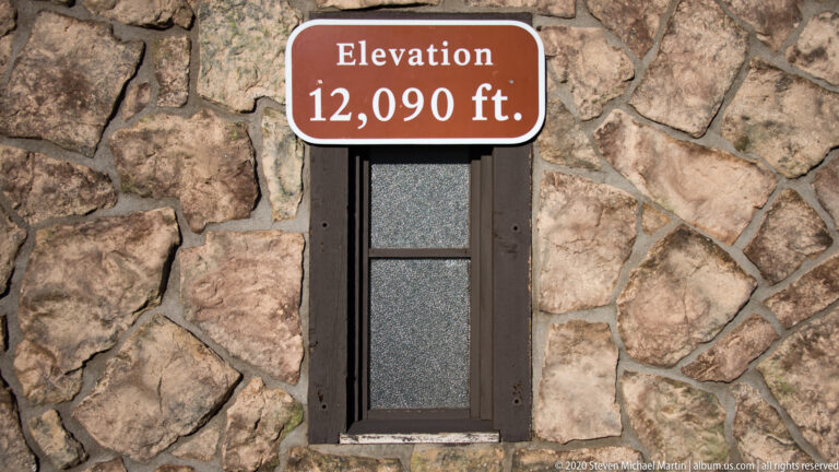 Red-brown sign reading “Elevation 12,090 ft.” is mounted on a rough masonry wall.