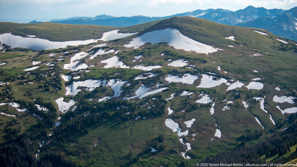 Patchy snow follows alpine basins and ridges above dark forests in a high mountain landscape.