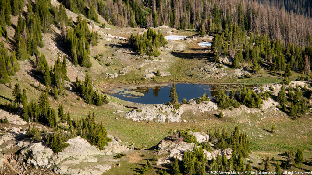 A small pond reflects conifers below a rocky alpine slope dotted with snow.